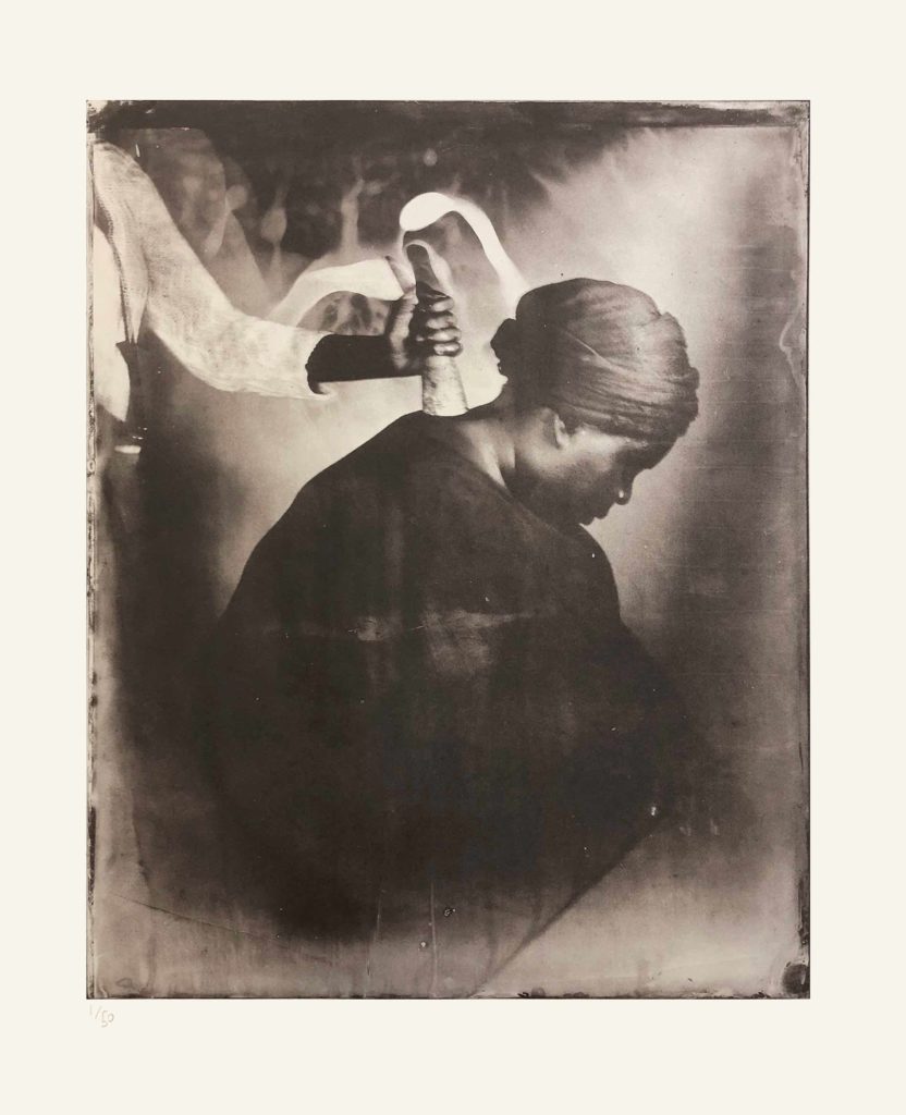 Alt-text: A person only partially visible places a cow horn on the back of Khadija Saye’s neck