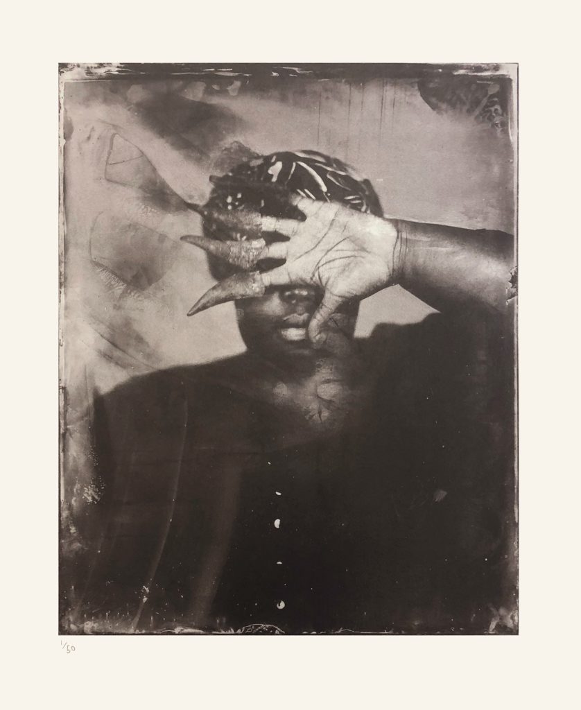 Alt-text: Khadija Saye’s hand, palm outward and with small goat horns on her thumb and fingers, obscures her face 